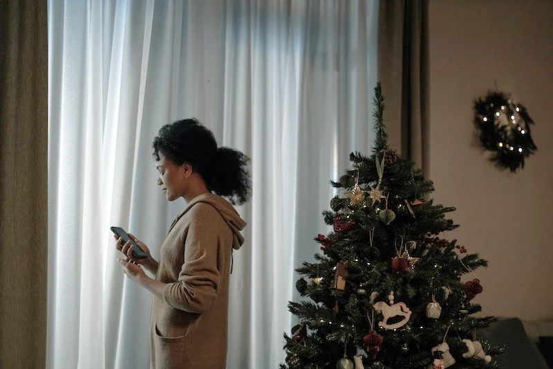 Image of a sad woman by Christmas tree, holding phone near window. Dealing with grief during the holidays.